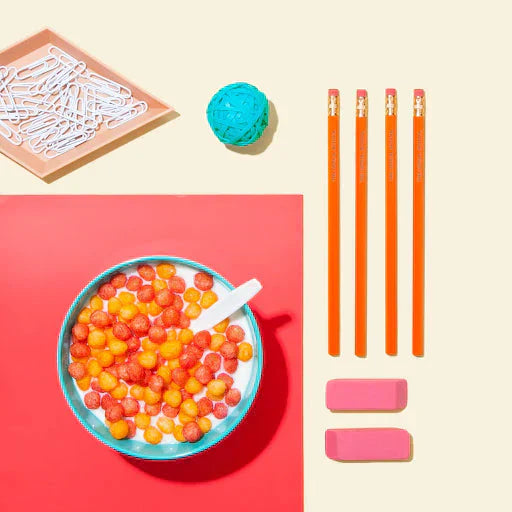 Bowl of colorful Schoolyard Snacks cereal with milk on a desk beside pencils, erasers, and paper clips.